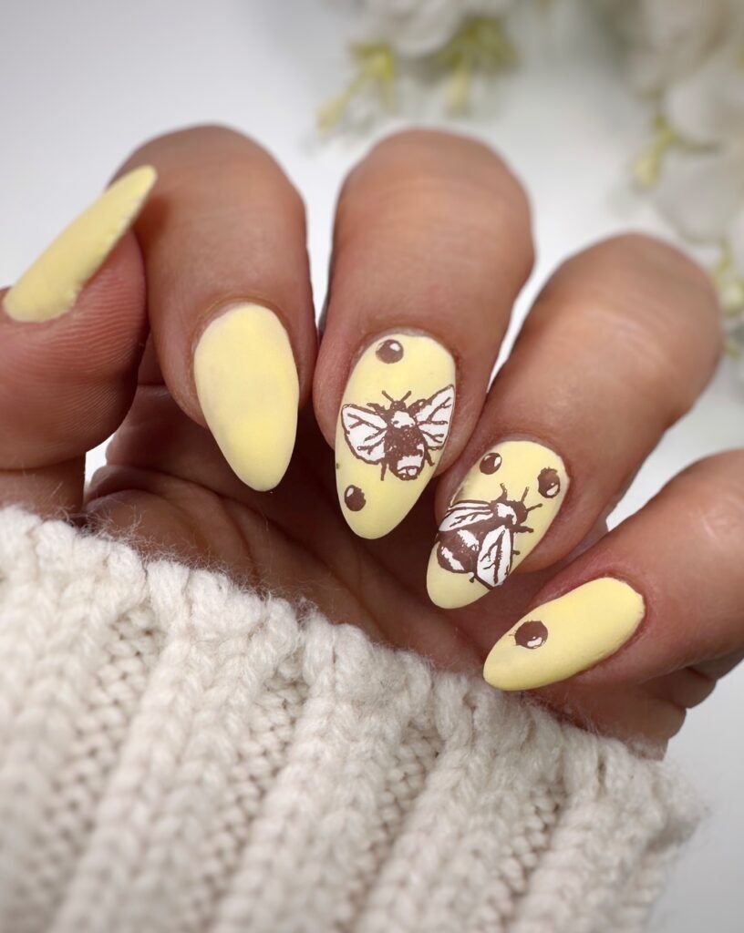 Yellow bee-themed nail art with black accents on almond-shaped nails. Cozy sweater background.