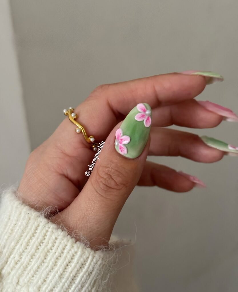 Hand with floral 3D nail art design, featuring green and pink colors, adorned with a gold ring and sweater sleeve.