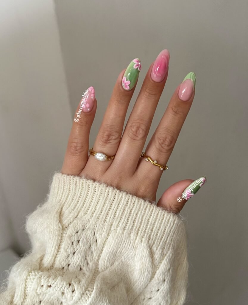 Hand with pink and green floral nail art, wearing gold and pearl rings, in a cozy cream sweater.