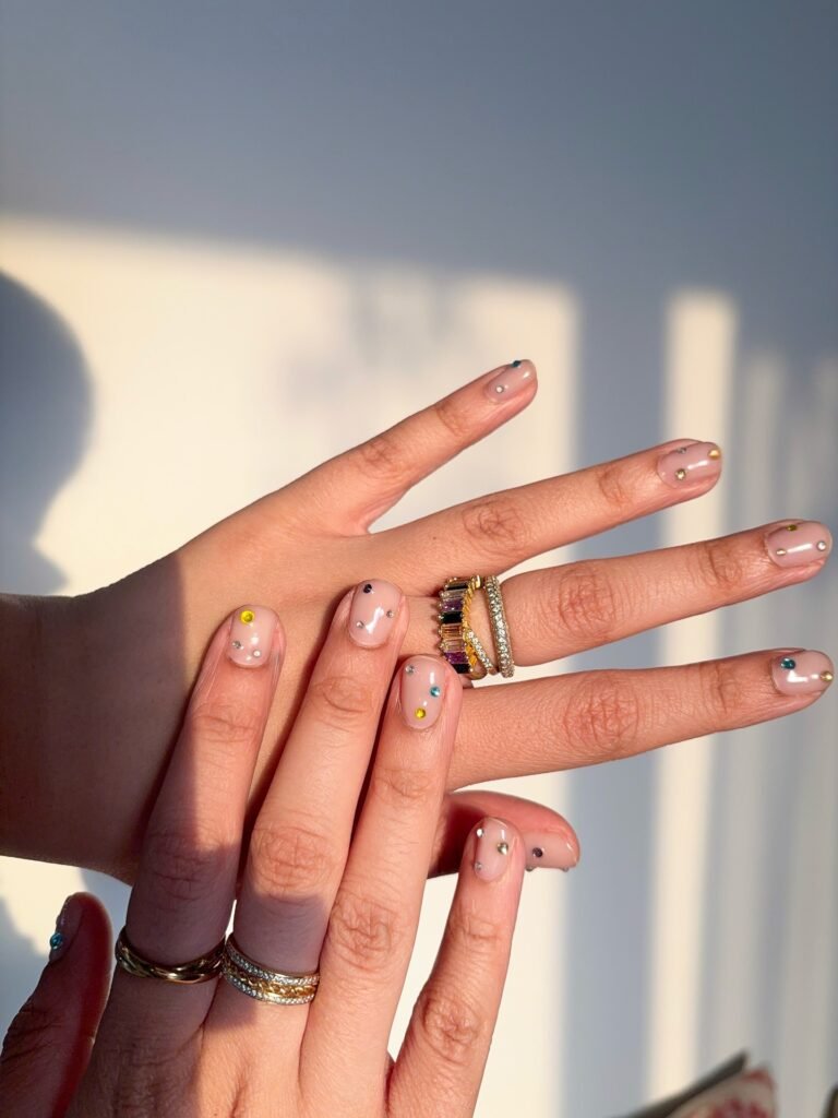 Stylish manicure with minimalist nail art and colorful dots, featuring gold and gemstone rings catching sunlight.