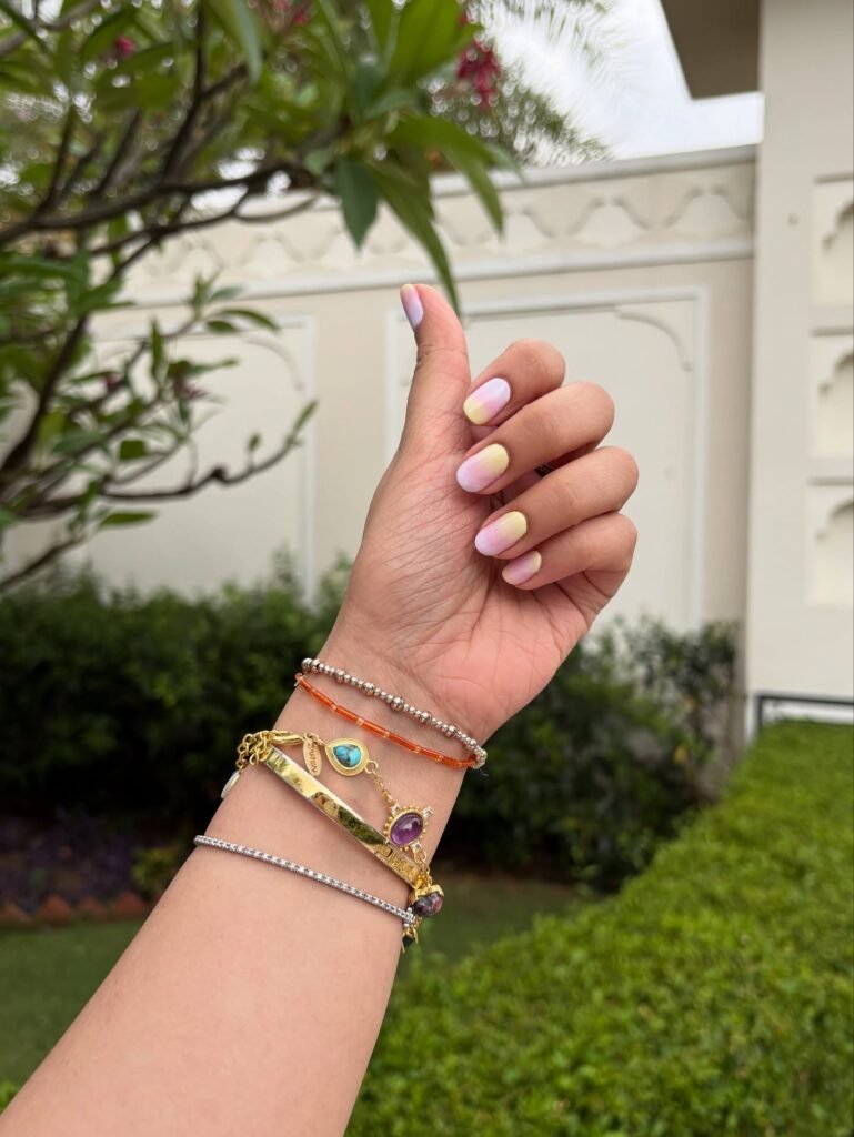 Colorful manicured nails and bracelets against a garden backdrop, showcasing vibrant jewelry and nail art style.