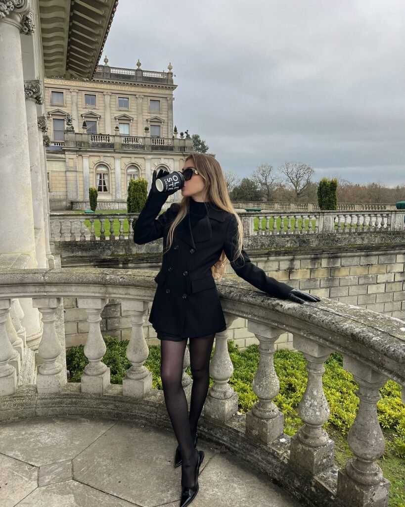 Woman in black outfit sipping from a mug on a balcony overlooking a historic building.
