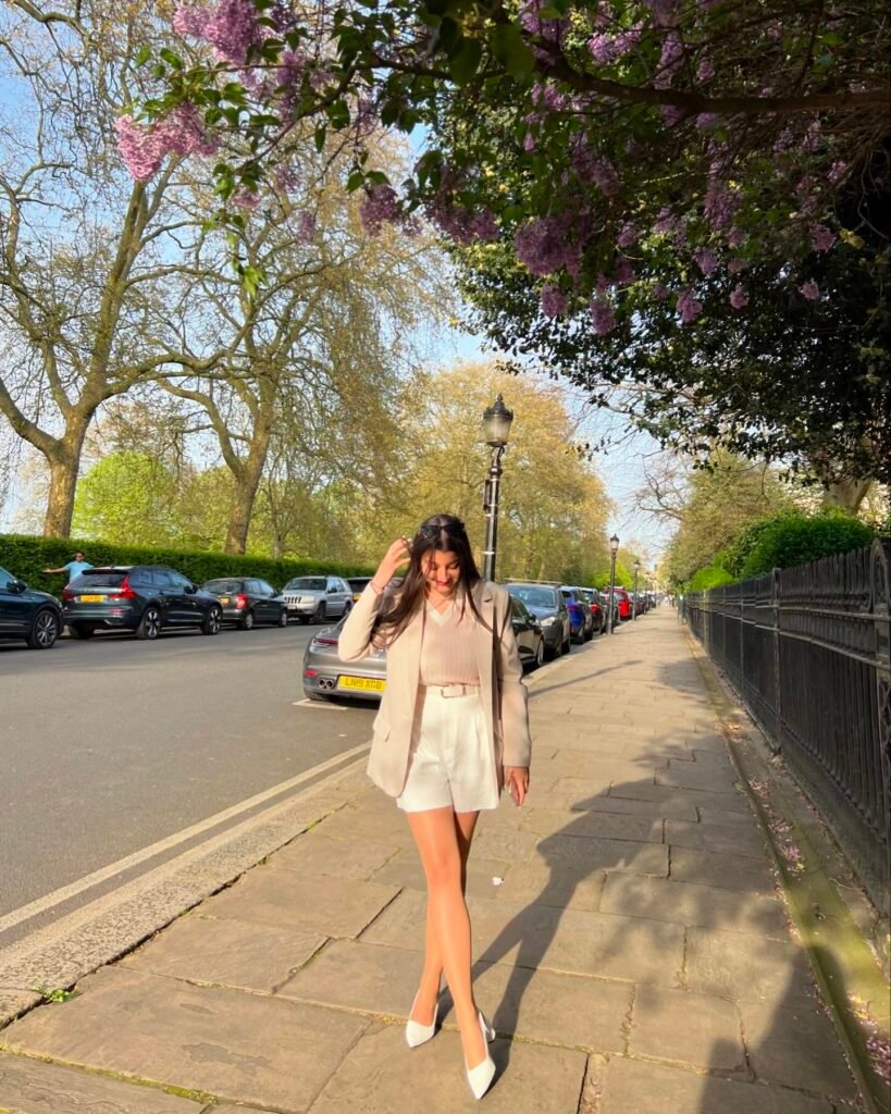 Woman in a stylish outfit walking on a sunny street lined with blooming trees and parked cars.