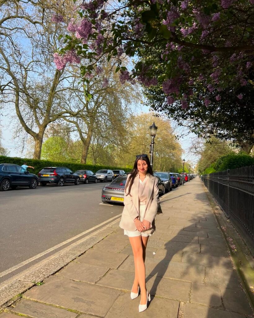 Woman in stylish outfit standing on sunlit sidewalk, surrounded by trees and parked cars on a bright day.