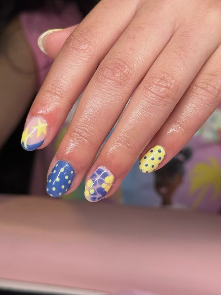 Colorful nail art with star, polka dot, and floral designs in blue, yellow, and purple hues on manicured nails.