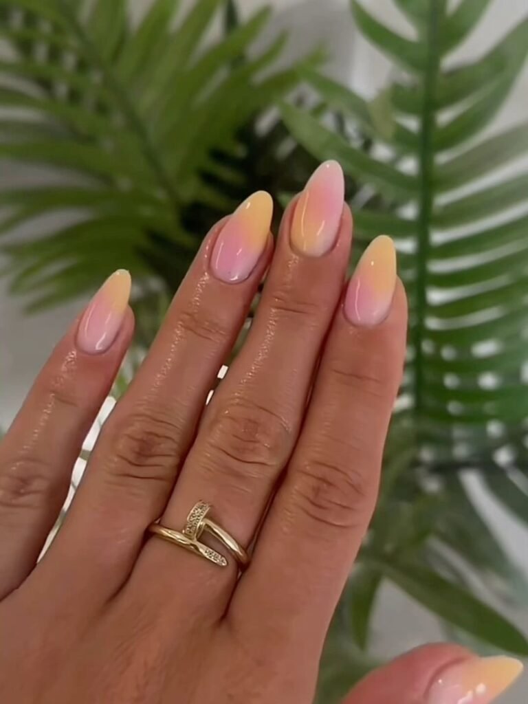 Gradient pink and orange stiletto nails with a gold ring, against a green plant background.