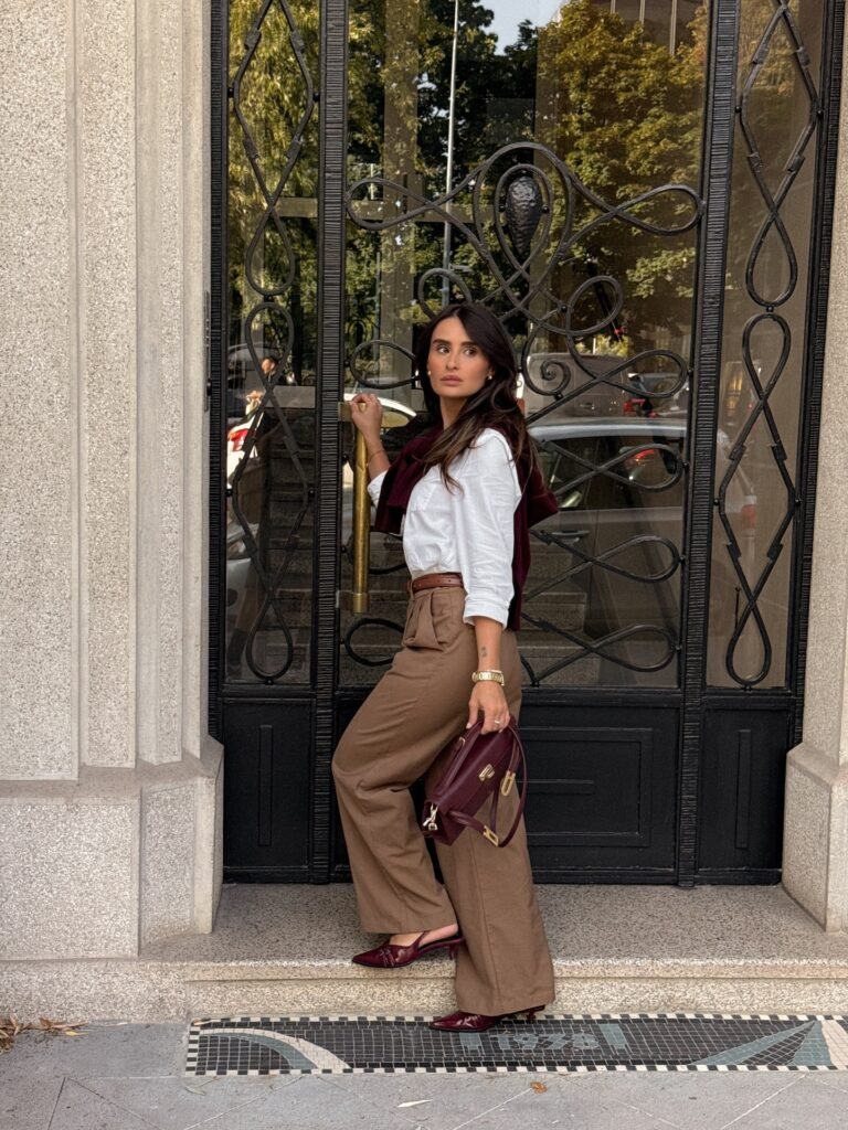 Fashionable woman in a white shirt and brown trousers holding a burgundy handbag, standing by an ornate door.