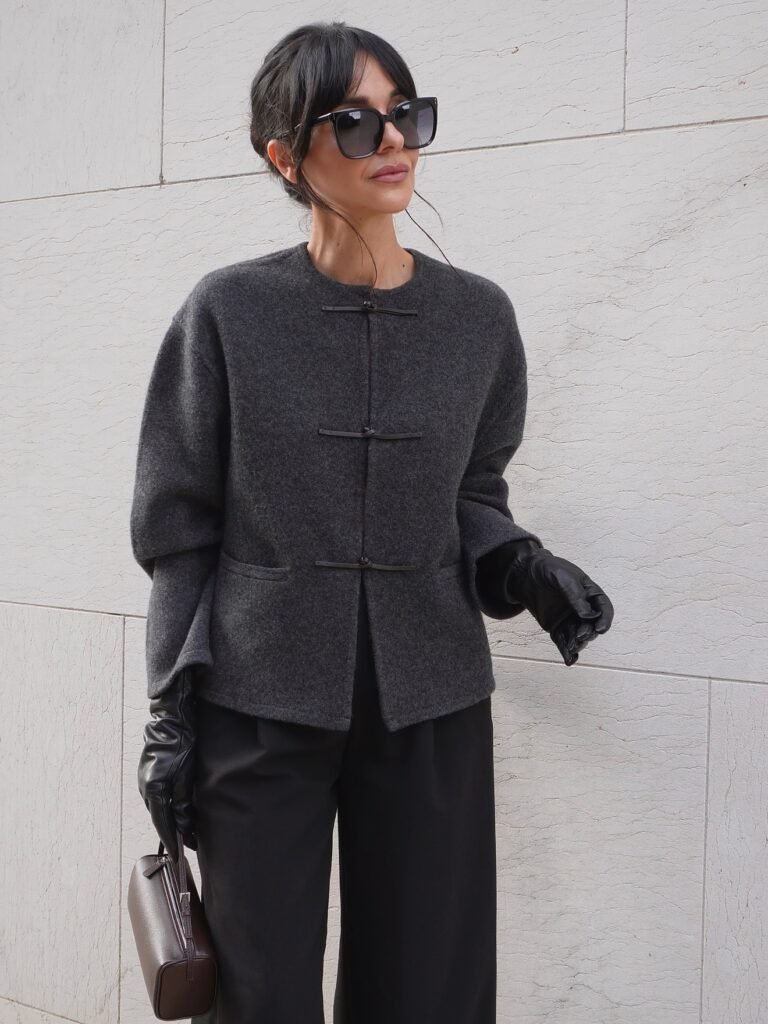 Chic woman in a stylish gray coat, sunglasses, and gloves holding a brown purse against a minimalist wall background.