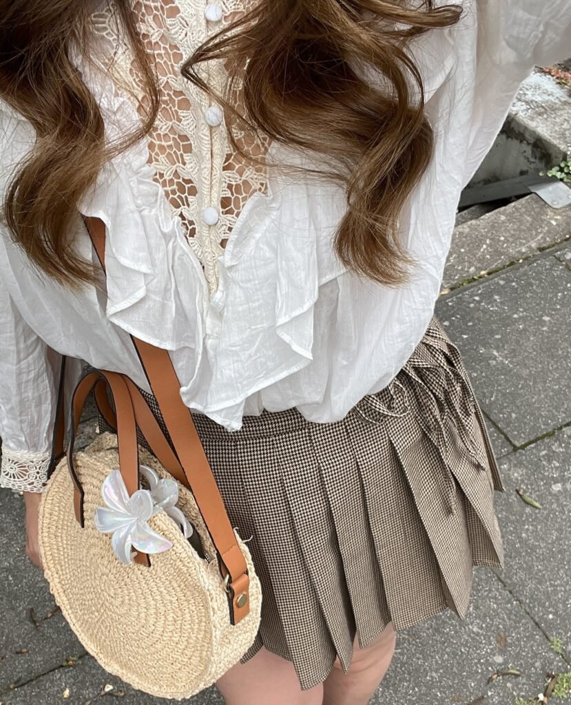 Woman in a stylish white lace blouse with a plaid skirt and straw handbag on a city street.