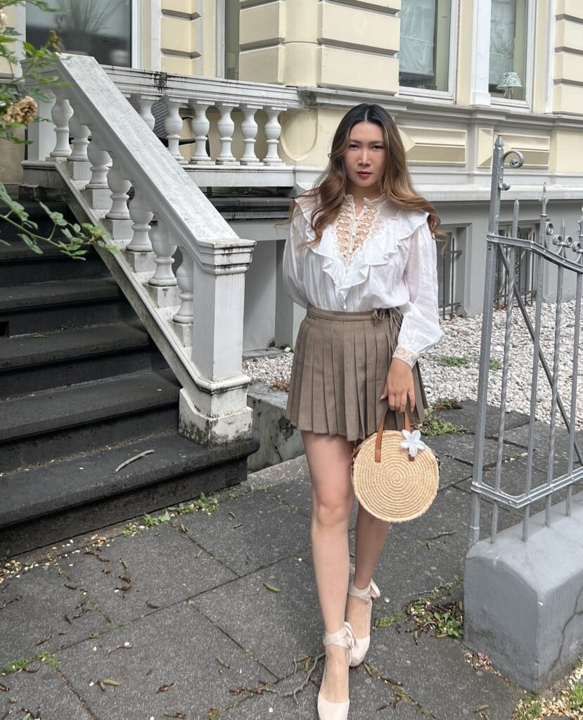 Woman in white blouse and brown skirt holding a wicker bag, standing on a stylish urban sidewalk.