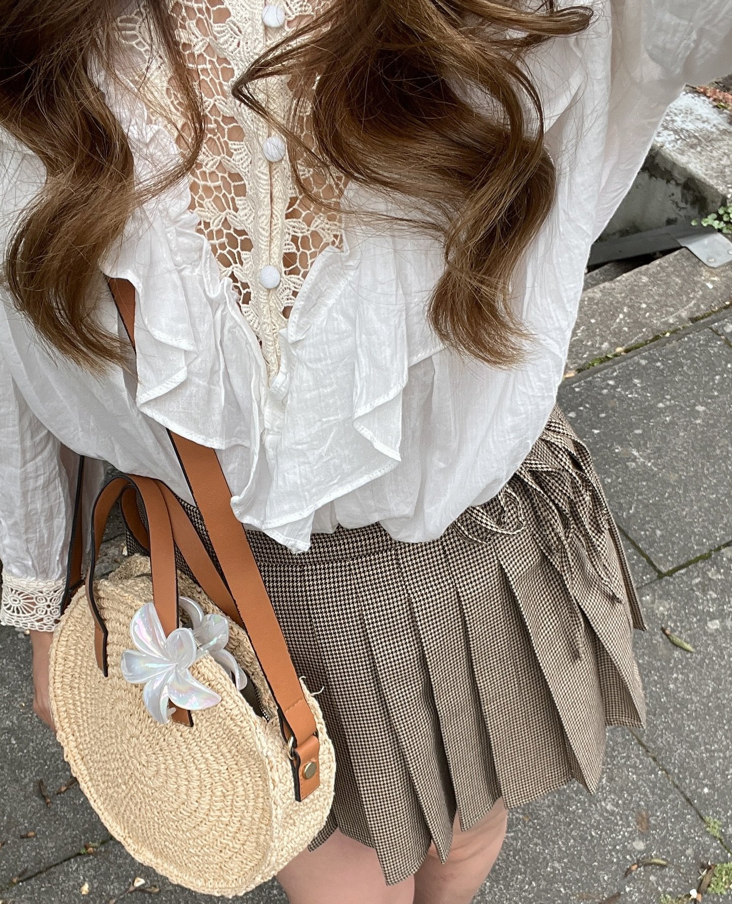 Woman in a stylish white lace blouse with a plaid skirt and straw handbag on a city street.