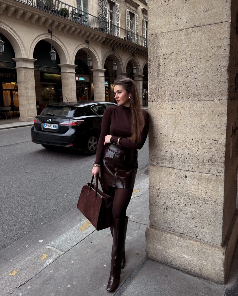 Woman in chic brown outfit leans against a city building, holding a matching handbag and wearing high boots.