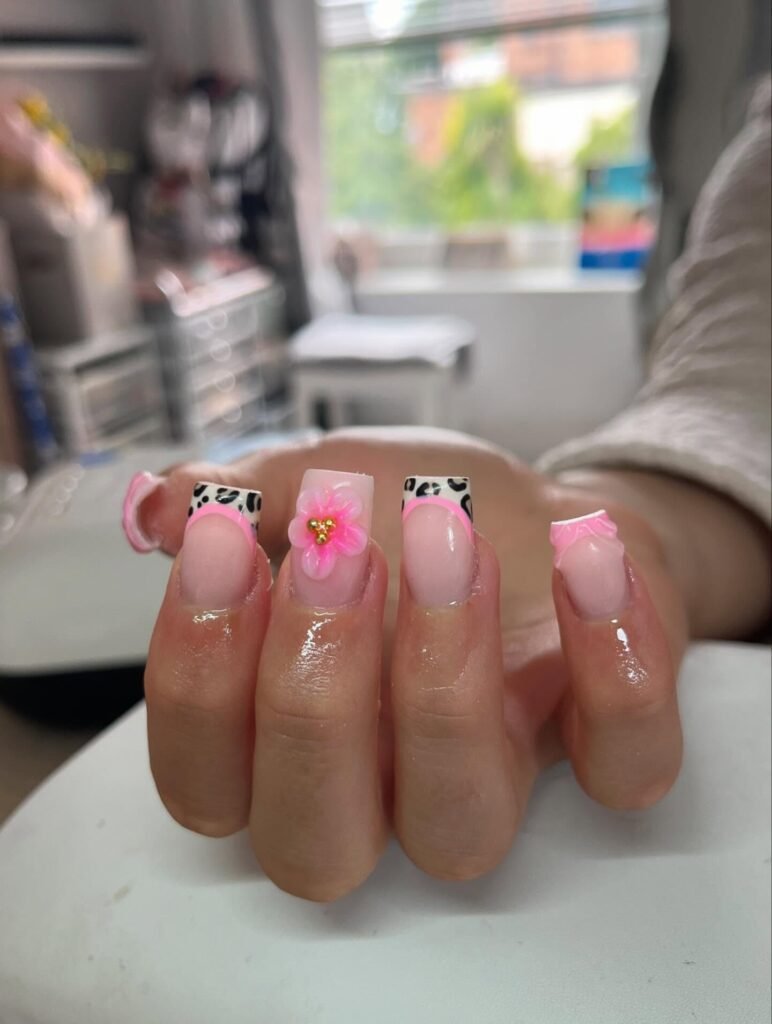 Pink and cow print acrylic nails with 3D flower accents, held in a bright, modern salon setting.