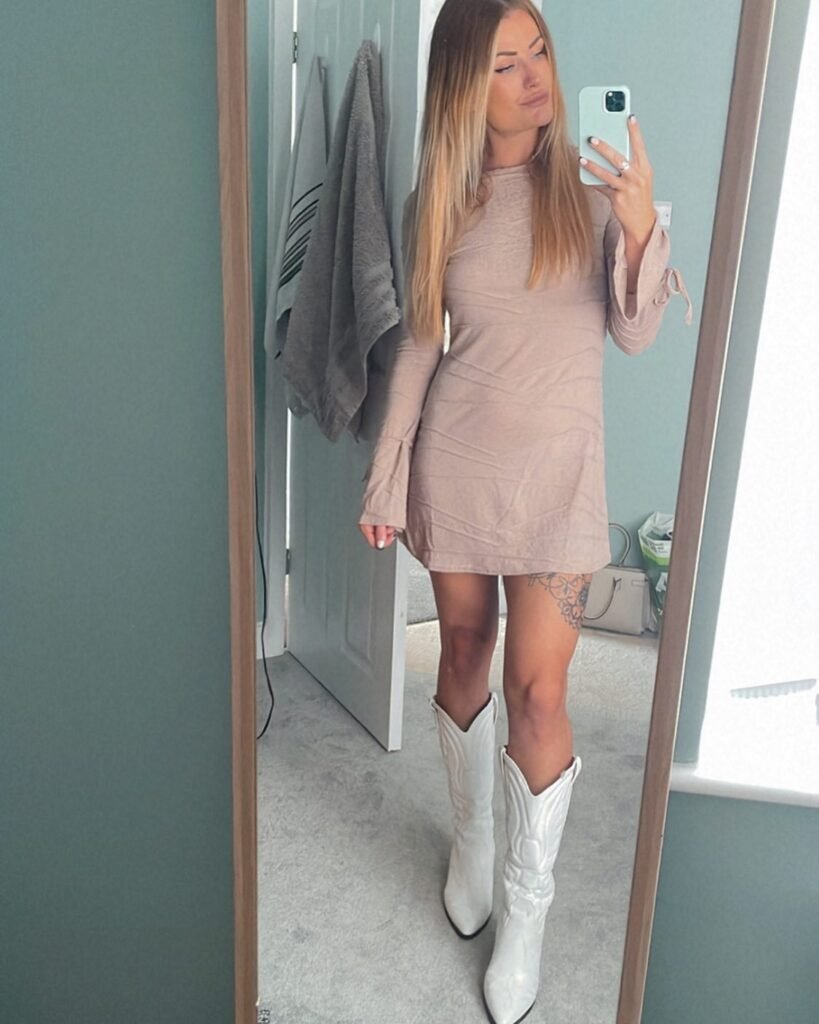 Woman in beige dress with white cowboy boots takes a mirror selfie in minimalist room.