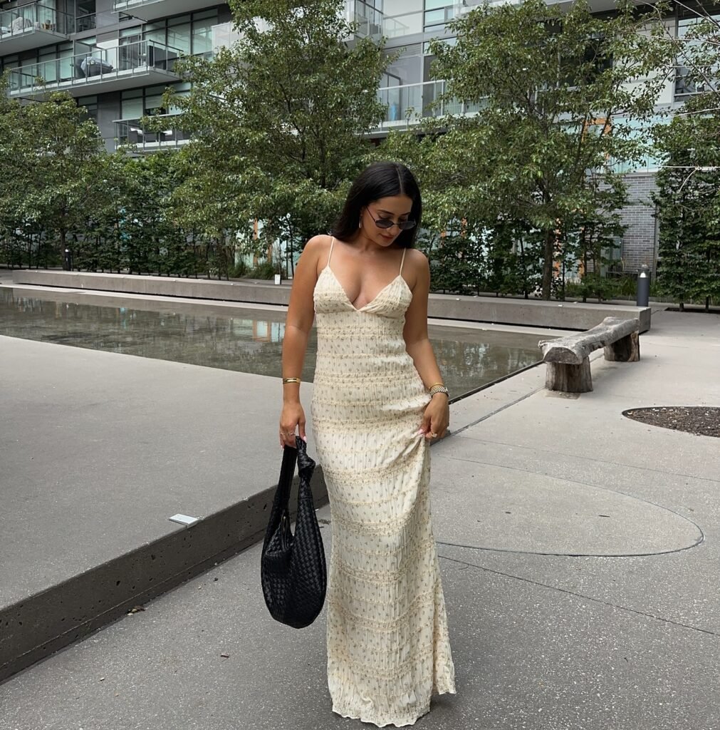 Woman in a cream dress with a black bag, walking outdoors near modern buildings and greenery.