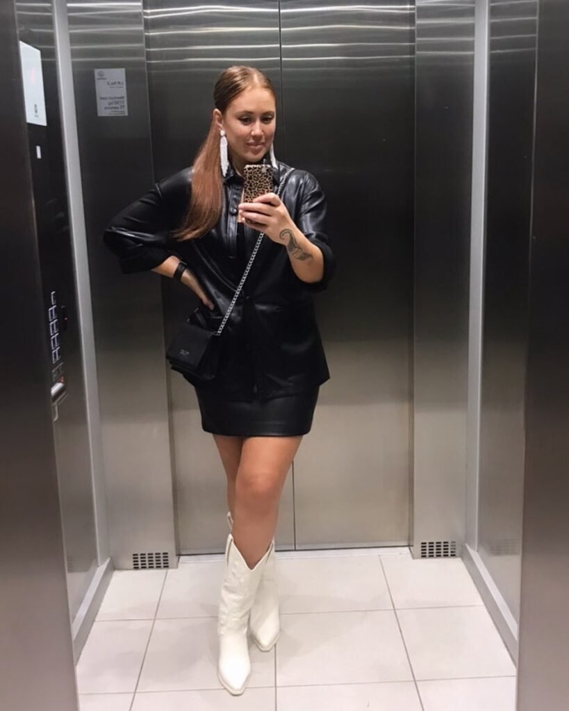 Woman in elevator wearing a black leather outfit, white boots, and long earrings, taking a mirror selfie.