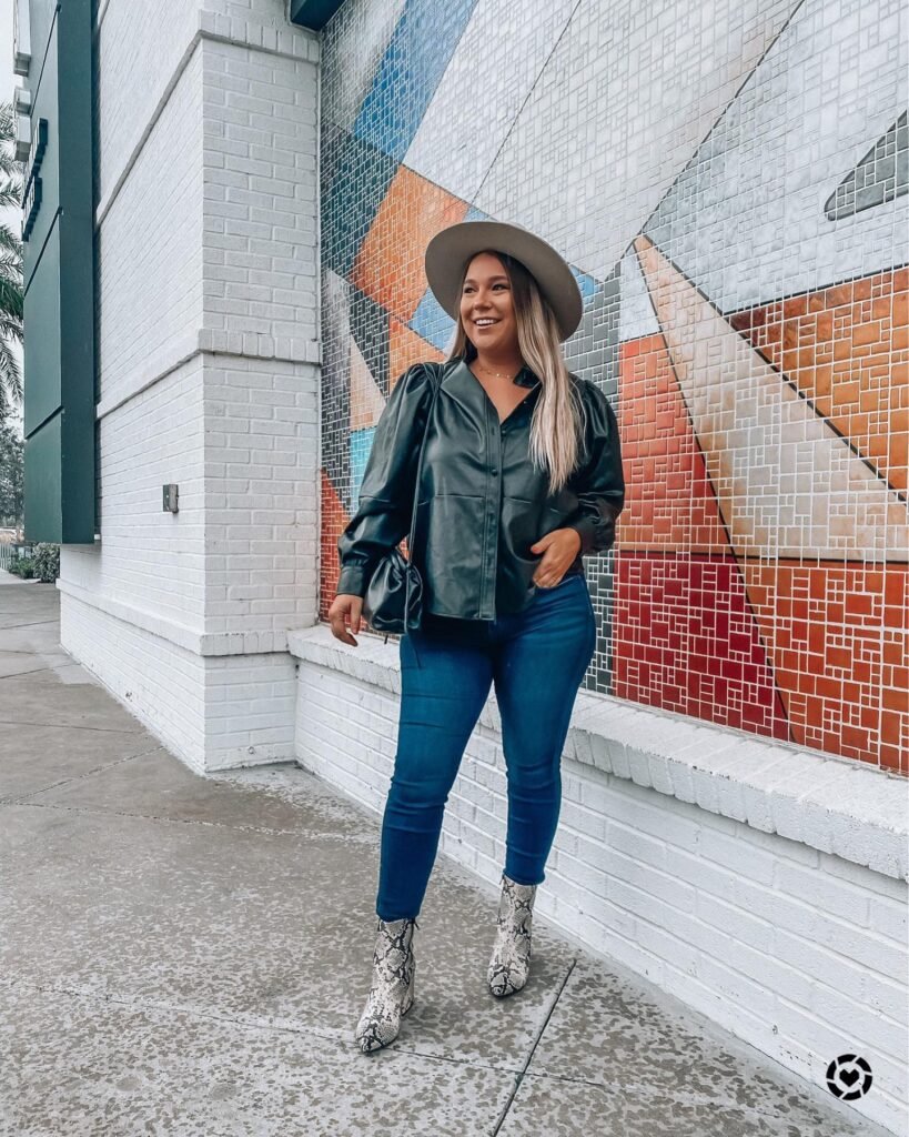 Smiling woman in hat, leather jacket, and jeans against colorful tiled wall.