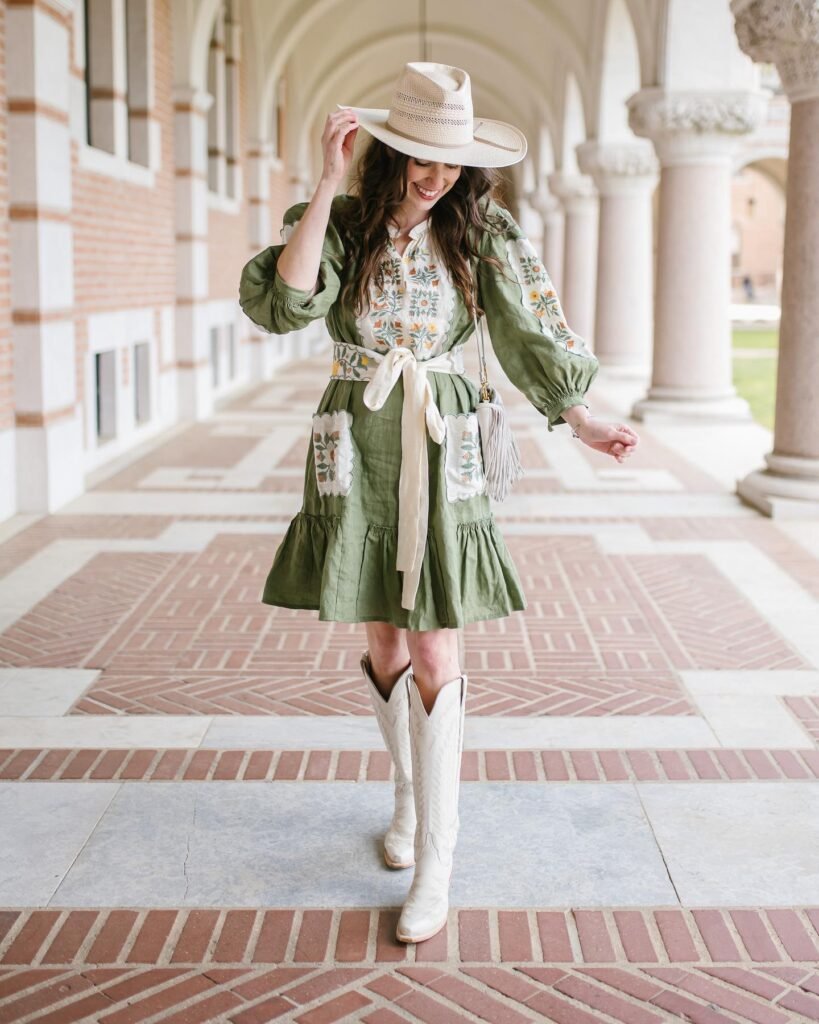 Woman in green embroidered dress and cowboy boots, smiling under arches. Southwestern fashion style.