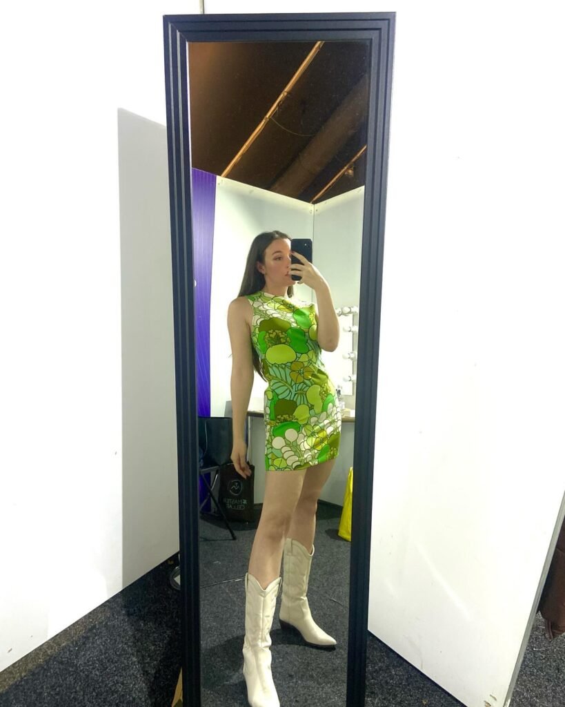 Woman in green floral dress and white boots takes mirror selfie, showcasing a chic, fashionable outfit.