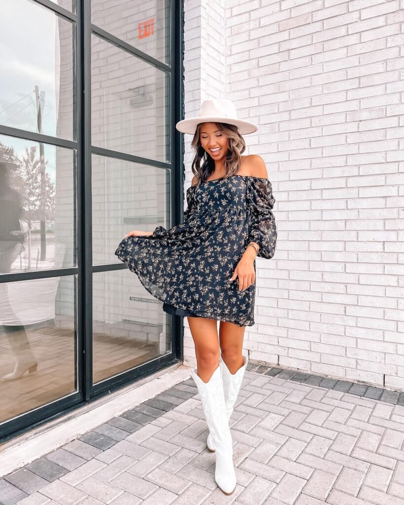 Woman in a floral dress and white boots poses by a window with a brick wall background. Fashion and style photography.