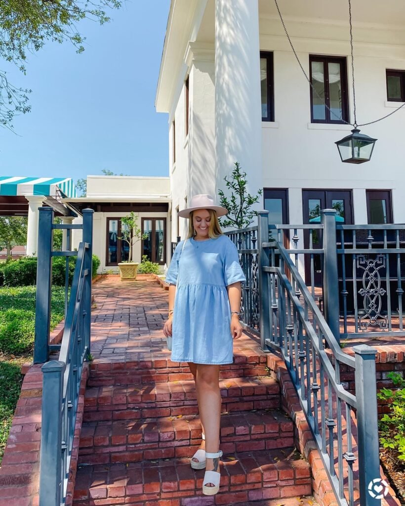 Woman in blue dress and hat standing on brick stairs outside a white building.