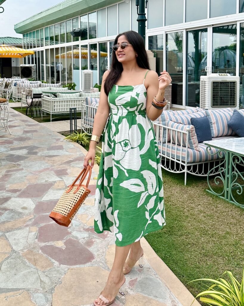 Woman in green floral dress and sunglasses walking outdoors, carrying a woven bag, with glass building in the background.