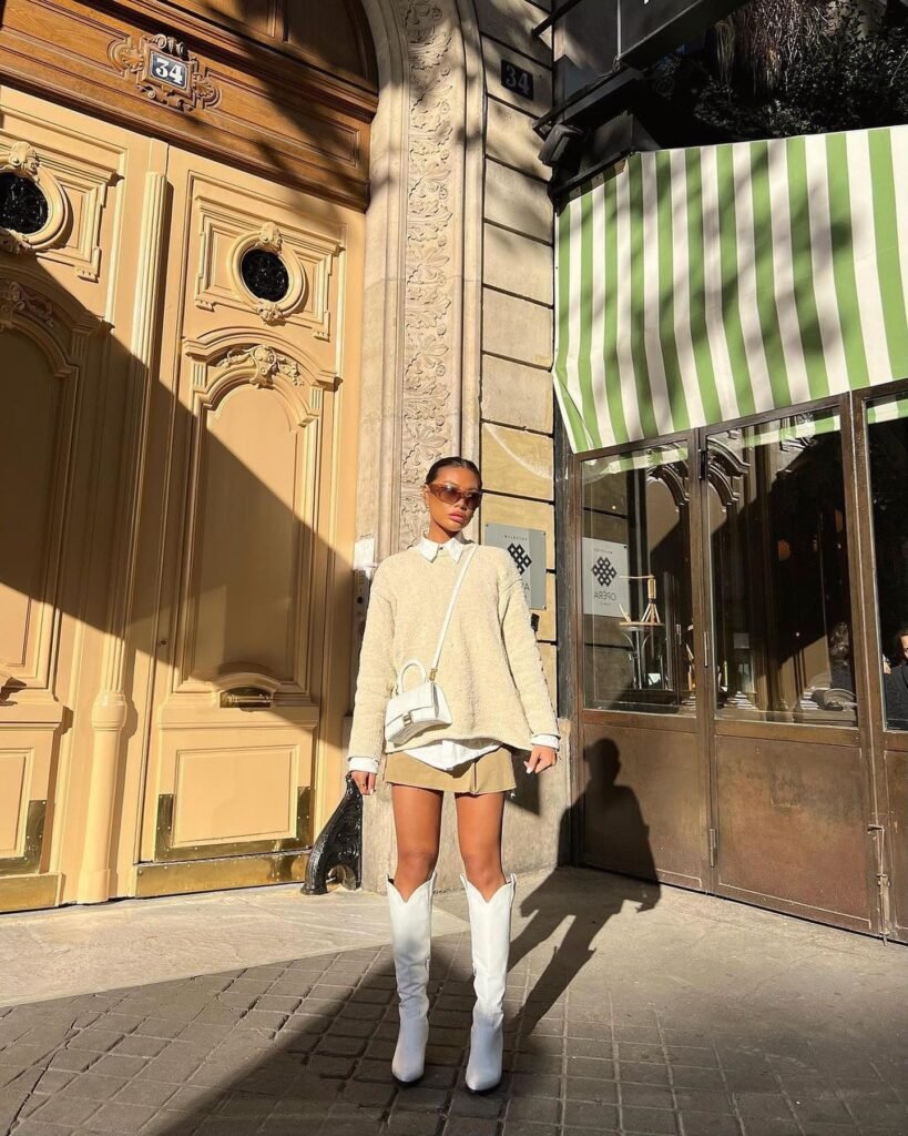 Fashionable woman in sweater, mini skirt, and white boots outside ornate wooden doors in sunlight.