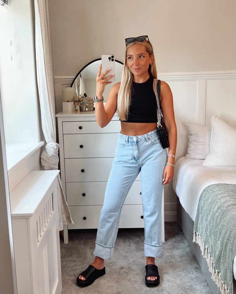 Woman taking a mirror selfie wearing a black top and jeans in a stylish bedroom with modern decor.