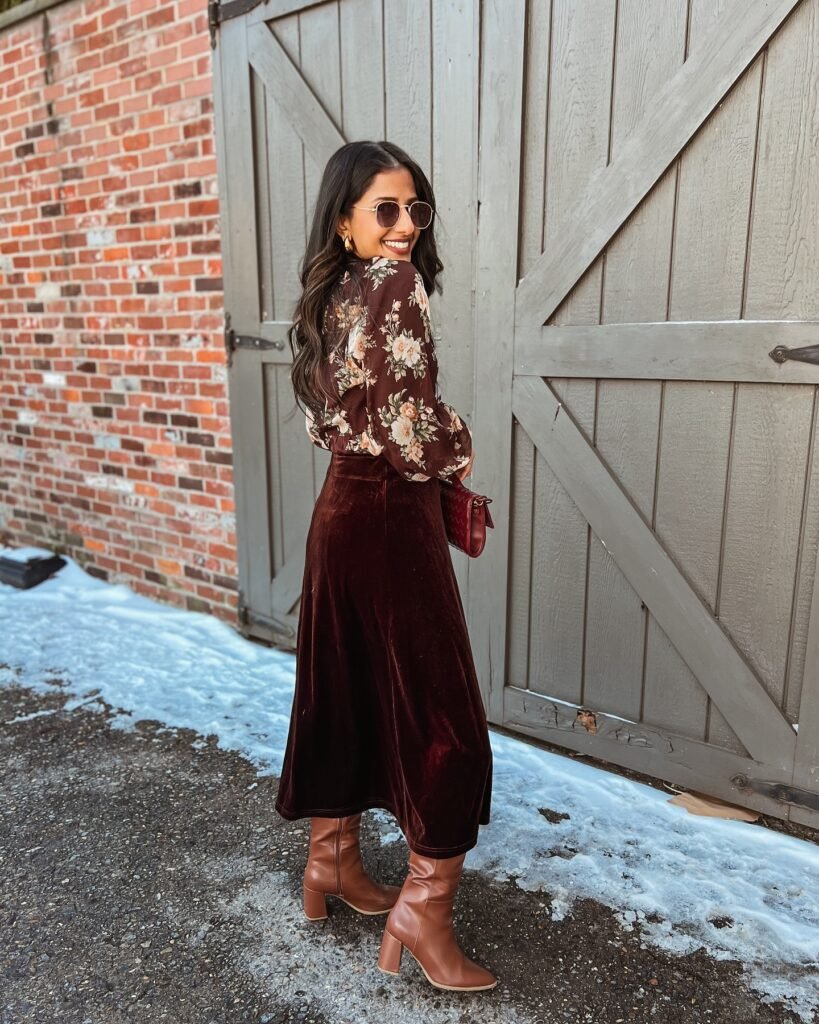 Stylish woman in floral blouse and maroon skirt, wearing sunglasses near a wooden gate.