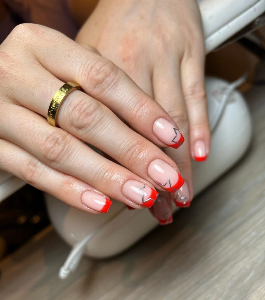 Red French tip nails with geometric silver accents, showcasing a stylish manicure and a gold ring on the finger.