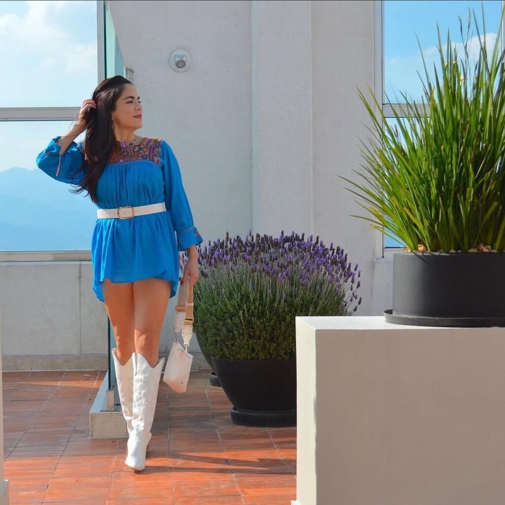 Woman in blue dress with white boots poses on a sunny terrace next to plants and lavender.