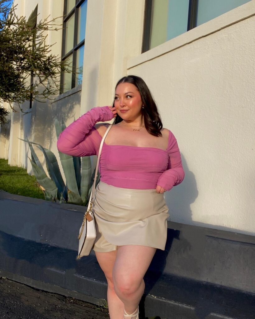 Woman in pink top and beige skirt smiles outdoors, holding a white purse in sunny setting. Fashionable spring look.