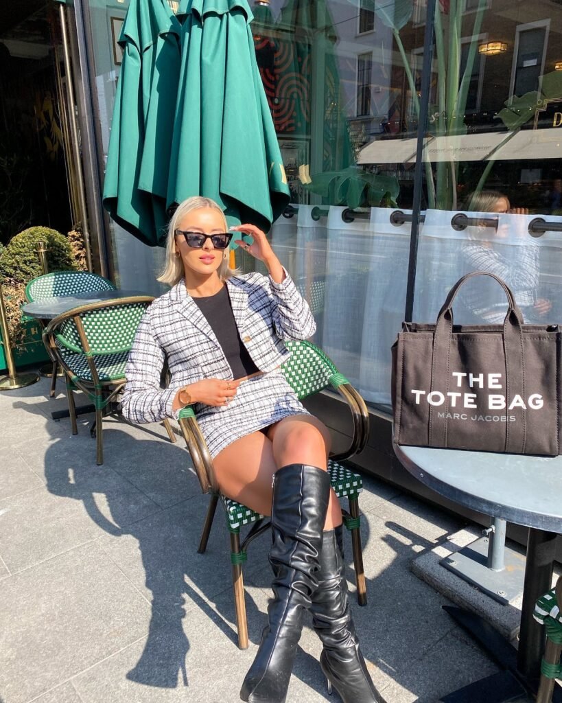 Fashionable woman in plaid outfit and sunglasses sitting at cafe with a designer tote bag and green umbrellas in background.