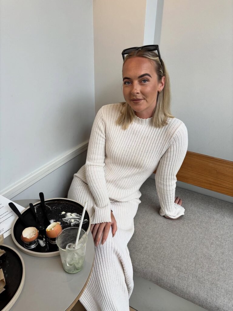 Woman in a white sweater dress sitting in a cafe, next to a table with breakfast remnants and a drink.