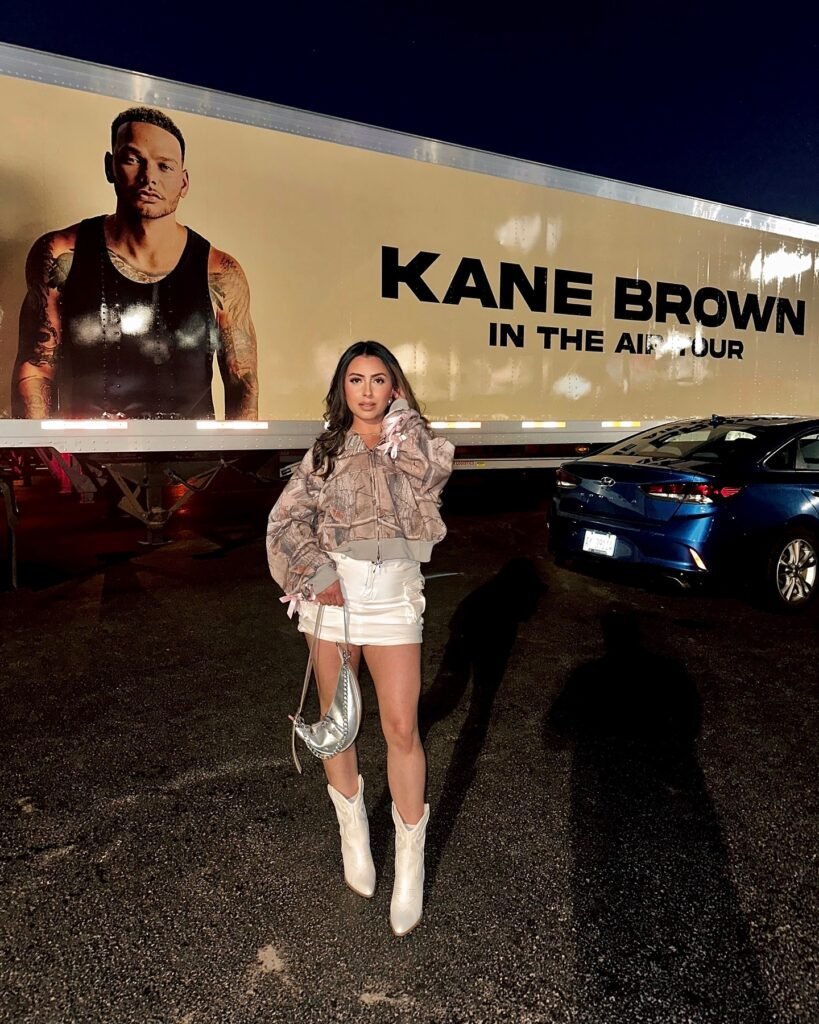 Person posing in front of Kane Brown tour truck at night in stylish outfit, holding a silver bag, wearing white boots.