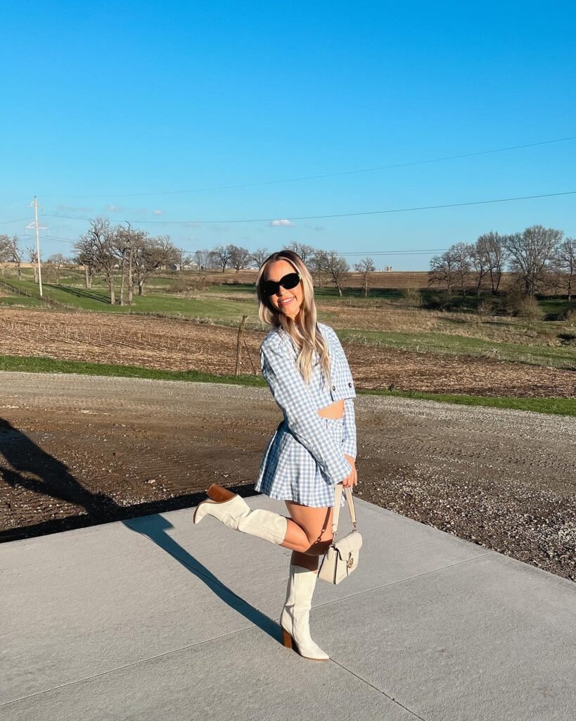 Woman poses outdoors in a blue checkered dress and white boots on a sunny day, holding a beige handbag.
