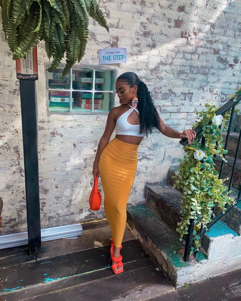 Fashionable woman in chic outfit posing by stairs with plants, wearing yellow skirt, white top, and holding orange handbag.