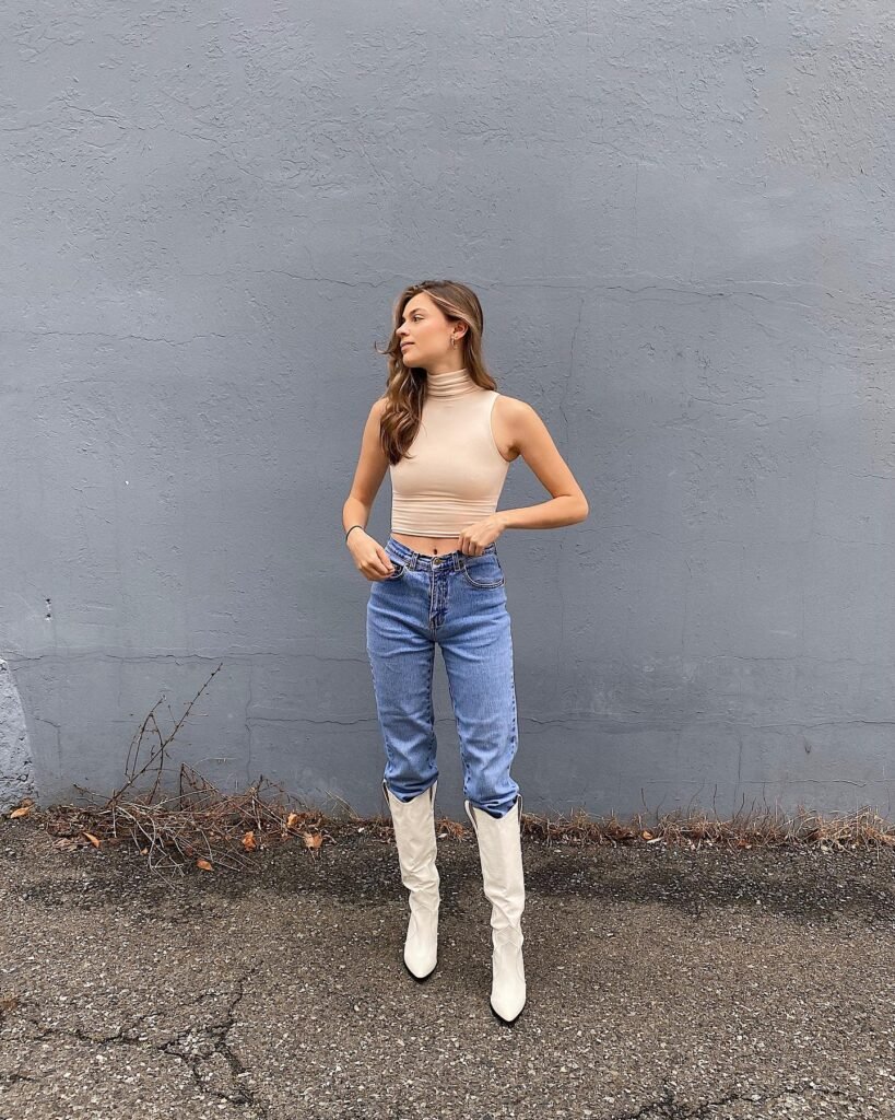 Woman in beige top with jeans and white boots stands against a gray wall.