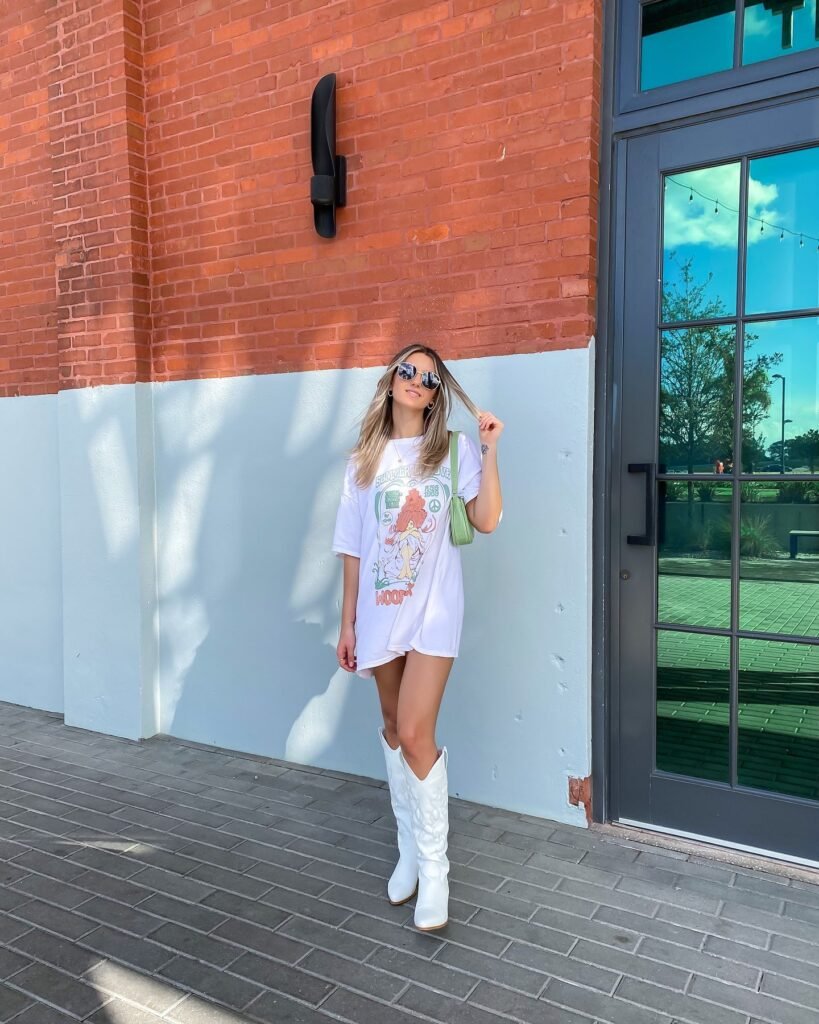 Woman in white graphic tee and cowboy boots poses by a brick wall and glass door.