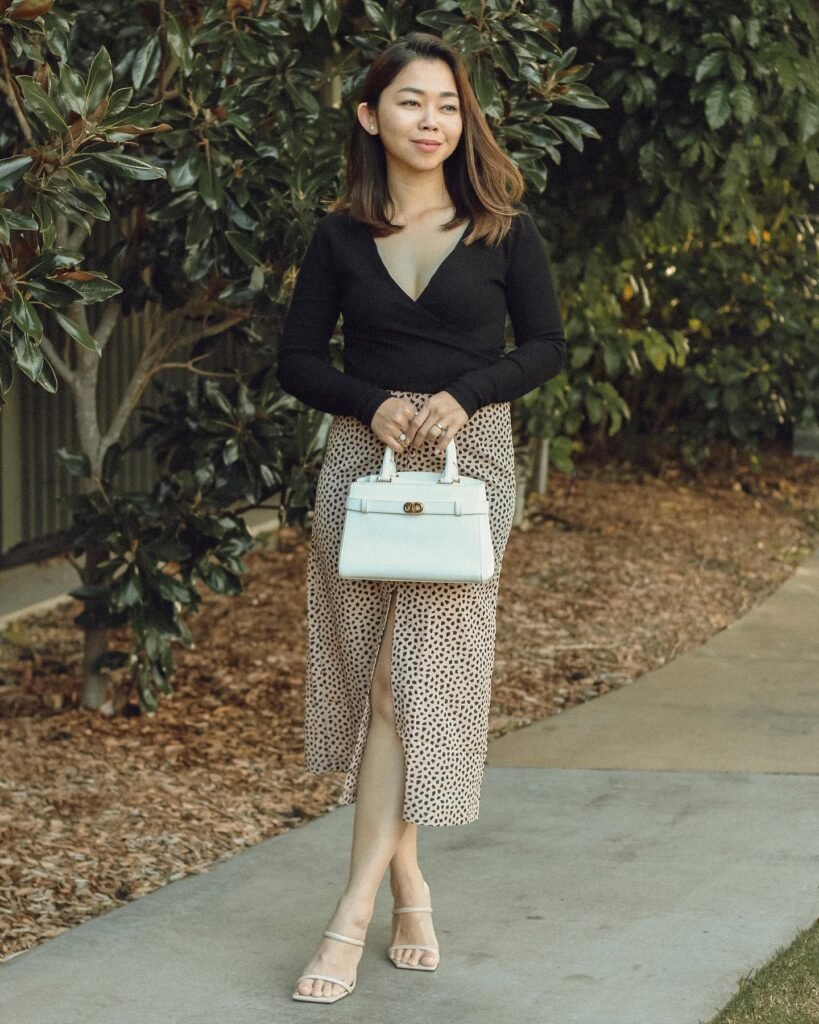 Woman in black top and patterned skirt walks on pathway holding a white handbag.
