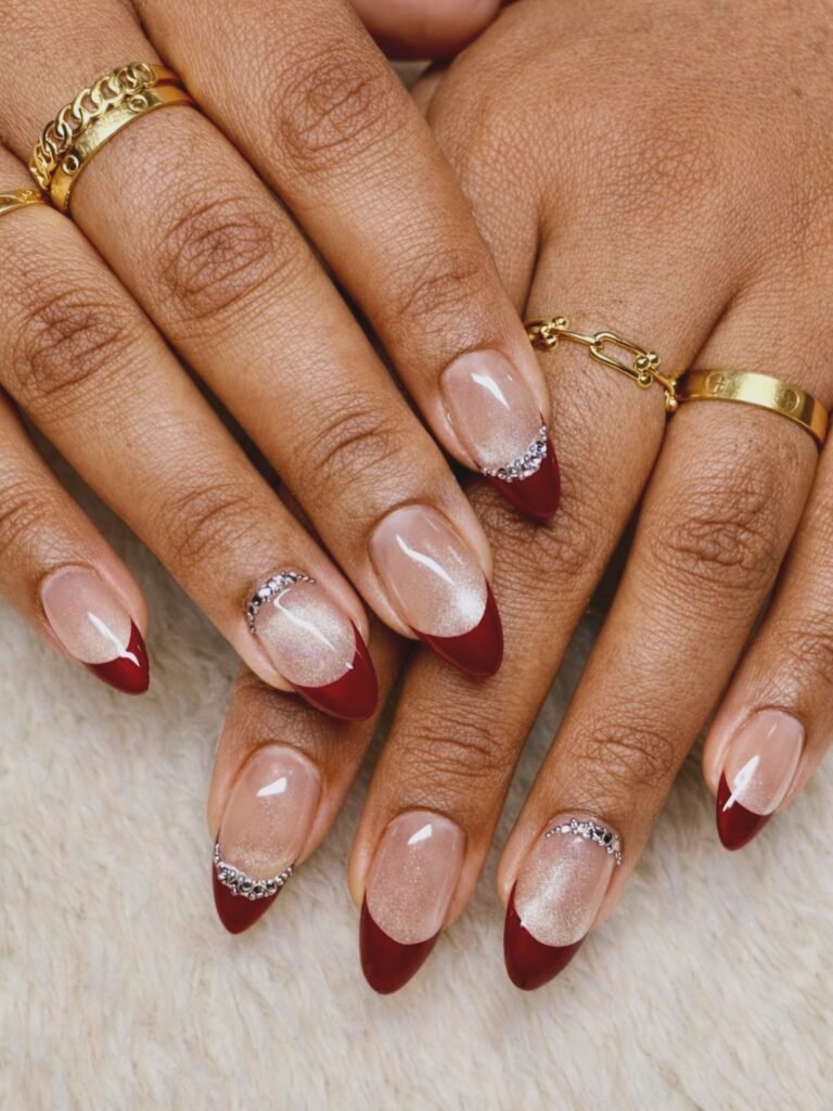 Close-up of elegant red-tipped nails with silver accents and gold rings on hands, against a soft background.