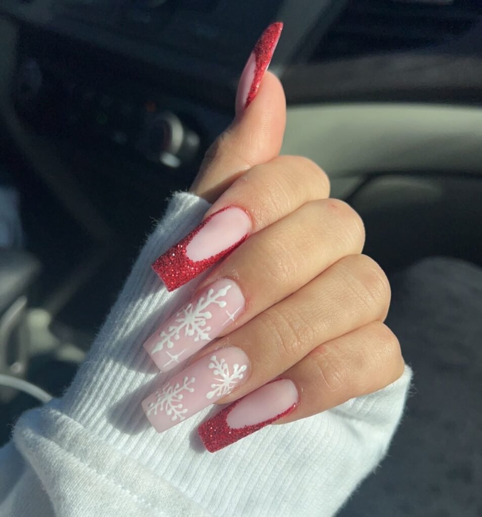 Festive nail art with red glitter tips and white snowflake designs on a cozy sweater background.