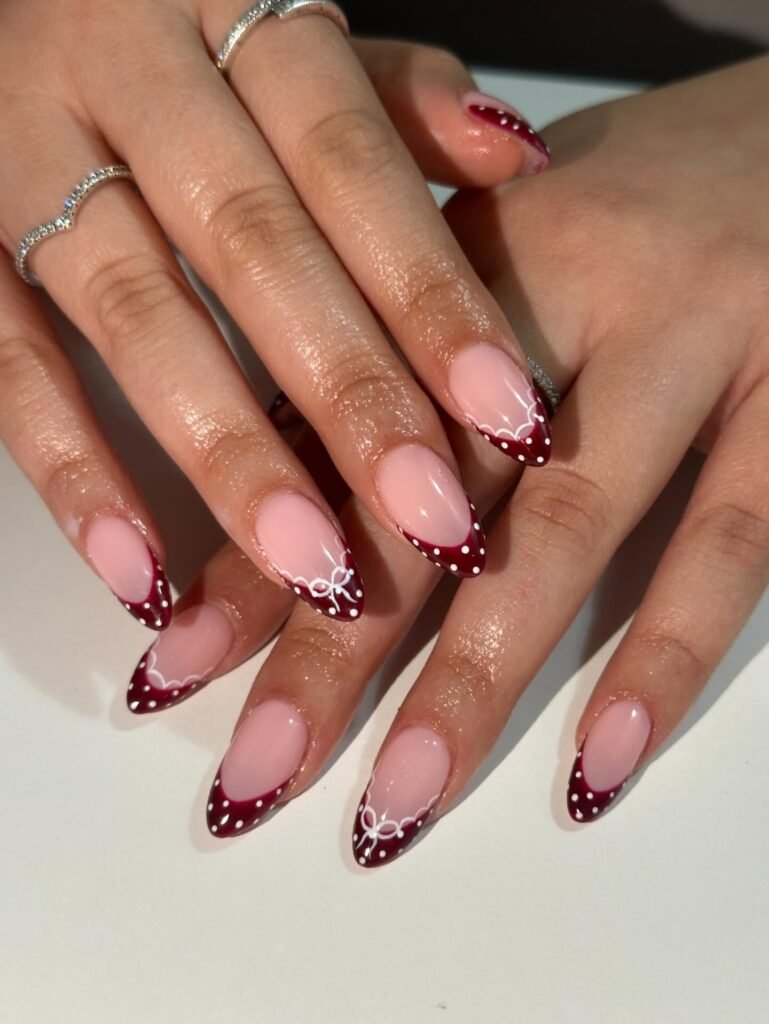 Elegant ombre nail design with burgundy and white polka dots, adorned with silver rings on well-manicured hands.