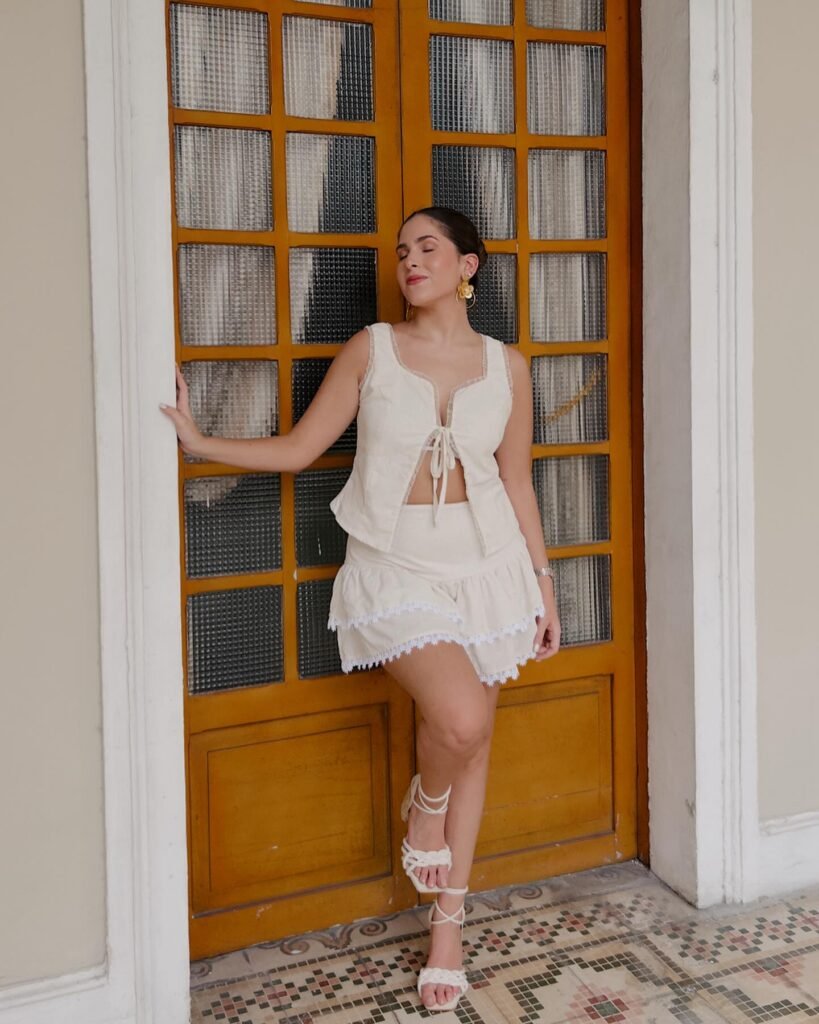 Woman in stylish white outfit and sandals posing by a wooden door, showcasing elegant fashion and relaxed style.