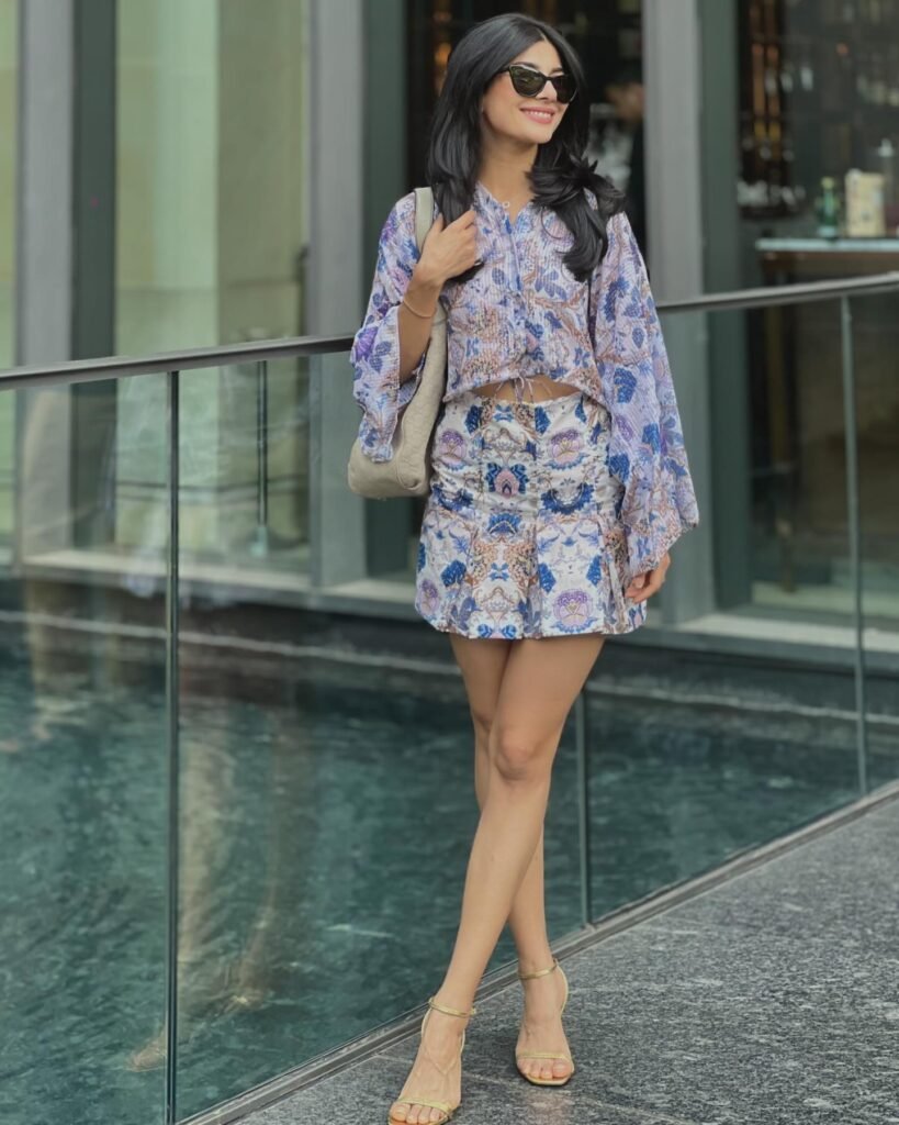 Woman in floral outfit and sunglasses standing by glass railing near water.