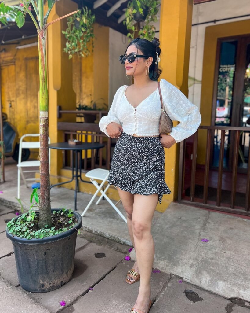 Woman in stylish outfit with sunglasses and purse poses outdoors near yellow wall and potted plant.