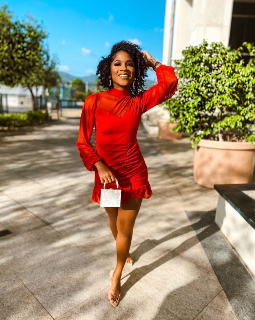 Woman in a red dress smiling and walking outside on a sunny day, holding a small white handbag.