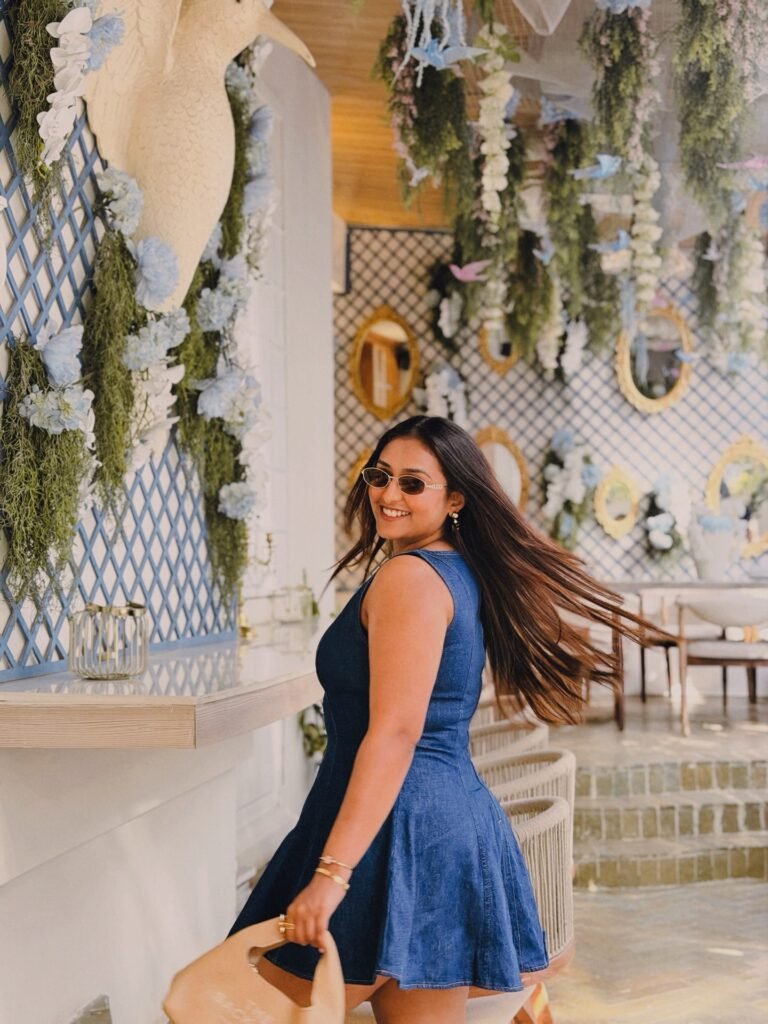 Woman in a blue dress enjoys a stylish, floral-decorated cafe with mirrors, greenery, and elegant decor.