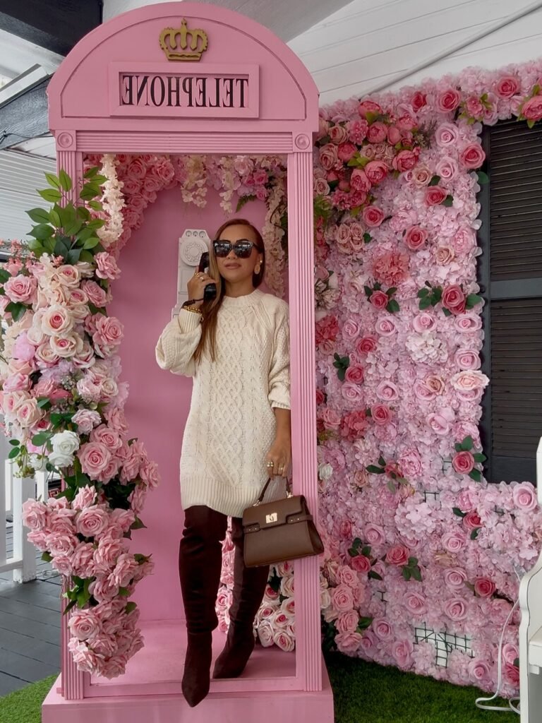 Woman in stylish outfit poses in pink floral phone booth with roses.