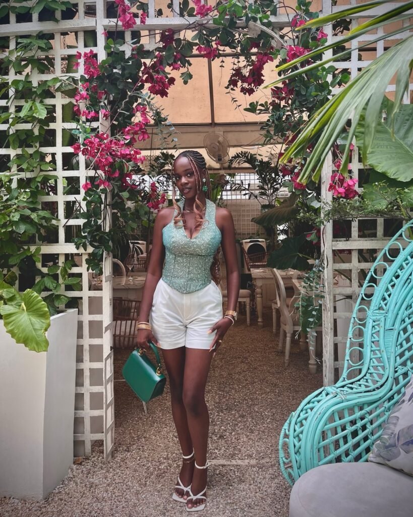 Woman in teal top and white shorts, standing under floral arch in garden setting, holding a green purse.